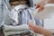 Close up of female hands pouring liquid laundry detergent into cap. Washer machine and clothes with wicker basket in background