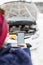 Close up of female holding blank screen smart phone and roadside assistance insurance membership card against broken car. Winter