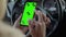 Close-up of a female phone hand with a green screen, the other hand on the steering wheel of a car.