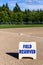 Close up of Field Reserved sign on empty local baseball field, third base and baseline, on a sunny day with woods and blue sky in