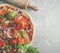 Close up of fresh pizza with tomatoes, onion, basil, cheese and salami and rolling pin on grey table with flour. Cooking