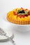 Close up of a German Fruit Flan topped with peaches, strawberries and kiwi, against a light background.