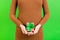 Close up of gift box in pregnant woman`s hands against her abdomen at green background. Young mother in brown dress. Happy