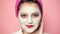 Close up of a girl with a face mask eats an apple on a pink background. Funny beautiful girl with cosmetic mask on her