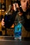 Close-up on glass with ice cubes into which bartender pours blue beverage from cocktail jigger.