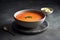 Close-up of a gray clay plate with fresh tomato soup and herbs. A spoon and a bowl of cream are next to each other on a dark