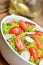 Close-up of green salad Lollo Biondo with tomatoes and radishes in a white bowl on wooden table, a pitcher of oil in the