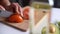 Close-up of hand of female cutting fresh tomato using kitchen knife on board.