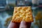 Close-up of a hand holding a golden cheese cracker with blurred background