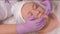 Close-up. The hands of a cosmetologist in lilac gloves apply a nourishing gel to a female face. A middle-aged woman is