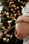 Close up of hands touching belly of young pregnant woman on a background of Christmas tree and lights. Expectation. Happy new year