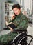 Close up of a handsome young soldier sitting on wheel chair reading a book at home and using his cellphone in a blurred