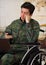 Close up of a handsome young soldier sitting on wheel chair using his cellphone while his computer is over his legs