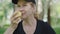 Close-up of happy Caucasian woman in sportswear and cap smelling tasty apple in summer park. Portrait of relaxed mid