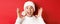 Close-up of happy young man in santa hat, making big christmas announcement, smiling amazed, standing over red