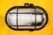 Close up image of kind of streetlight protected with black metal cage box. Set on bright yellow wall
