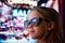Close-up image of little girl in sunglasses standing on street and looking at shop window. Lights reflection on glasses