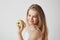 Close up of joyful small light-haired girl with blue eyes in white dress holding apple in hand, smiling brightfully in