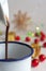 Close-up of jug serving chocolate over white cup with red Christmas decorations on bokeh, on white wooden background