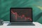 Close up of laptop on desktop with coffee cup, falling red business graph grid on chalkboard background. Crisis, recession and