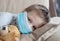 Close-up on a little boy in a medical mask on a sofa with a teddy bear. Pandemic, coronovirus, infection. Sick children