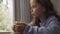 Close-up of a little grey-eyed girl with a cup of hot tea sitting at the table. Cute child in blue sweater enjoying hot