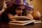 Close-up little witch, enchantress in wizard hat reading sorcery and spell magic book surrounded by Halloween atmosphere