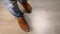 Close-up of a man in blue jeans and brown leather sneakers walking on a light-colored wooden floor. View from above