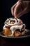 Close-up man\\\'s hand touching freshly baked cinnabon roll with sweet glaze on a black background. Generated AI