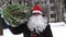 Close up of a man wearing a red Santa hat and a white beard carries home a Christmas tree packed in a grid