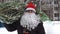 Close up of a man wearing a red Santa hat and a white beard carries home a Christmas tree packed in a grid