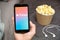 Close up mans hand holding a mobile phone with iTunes logo with Apple earphones and popcorn box next to him. Music