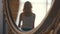 Close-up of mirror reflection of young blond woman in white t-shirt and blue jeans walking near a window in her