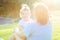 Close up mother and toddler baby daughter relaxing, embracing, laughing and have fun on green grass meadow at evening sunset. Spen