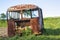 Close-up of old forsaken passenger bus with broken windows rusting in high green weedy grass on edge of plowed brown field on