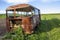 Close-up of old forsaken passenger bus with broken windows rusting in high green weedy grass on edge of plowed brown field on