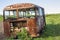 Close-up of old forsaken passenger bus with broken windows rusting in high green weedy grass on edge of plowed brown field on
