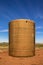 Close-up of old rusty water tank sitting in pasture with trampled red earth around it on flat ground with tree line in the