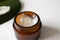 Close-up open cream in brown glass jar and tropical leaf on background on white, selective focus. Moisturizer butter for skin care