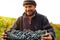 Close up photo of a farmer worker simple man in warm sweater and hat holding in his hands a box full with grapes for