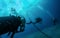 Close up photo of three divers swimming after each other close to anchor chain in blue water. Light at surface, air bubbles from