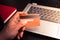 Close up photography of hand`s man holding a reverse credit card with the laptop behind on a wood table. Sunlight photography.