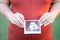 Close-up picture of pregnant woman in red maternity clothes holding ultrasound image of growing baby on her baby bump.