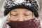 Close-up portait of a blonde woman in beanie hat and nose covered in scarf stands with eyes closed in winter snowfall. Nordic