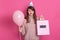 Close up portraiot of disappointed young girl in party hat and with trumpet and pink baloon, poses  over pink background,