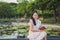 Close up portrait Asian woman in a white dress sitting next to a lotus pond, holding a book and looking at the camera in a public