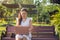 Close up portrait Asian woman in a white dress sitting and working with a pink laptop on a wooden bench in a public park