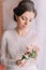 Close-up portrait of beautiful innocent bride in wedding dress near window holding cute boutonniere