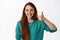 Close up portrait of beautiful young woman with red long hair, show thumb up and smiling pleased, looking at logo promo