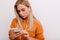 Close-up portrait of calm blonde female model with elegant make-up texting message on phone. Indoor photo of tired fair